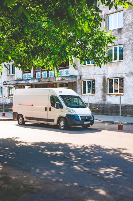 A white panel van parked on the street outside a residential building in the daytime, partially shaded by green foliage from an overhead tree. The building has multiple windows and a weathered exterio