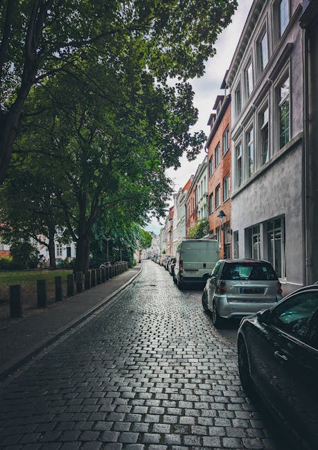 A narrow street lined with multi-storey residential buildings featuring large windows and brick facades in various colours, including grey, red, and white. The street is paved with cobblestones, and there are several parked cars, including a grey van positioned near a building's entrance, with others further down the street. A large tree with dense green foliage extends over the sidewalk, providing shade, and the overcast sky contributes to diffuse lighting. The scene captures a typical urban residential area suitable for house removals and furniture transport, illustrating the constraints of narrow streets and parking in Pentonville, which [COMPANY_NAME] navigates during home relocation and moving services, often involving loading and unloading furniture and packed boxes near the doorways and on the pavement.