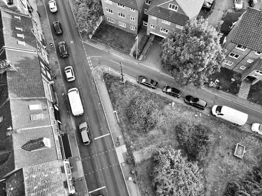 An aerial view of a narrow urban residential street with several parked cars lining both sides of the road, including a white van parked near the curb. Adjacent to the street are multi-story houses with brick exteriors, some with small porches and windows visible. A small grassy area with trees and bushes is situated next to the street, separated from the houses by a low pavement. The scene is captured in black and white, highlighting the tight-fitting parking and urban environment typical of a home relocation or furniture transport setting. The image reflects the logistics of moving services in confined city streets, with [COMPANY_NAME] operating within such environments to facilitate efficient packing, loading, and transportation of household items.
