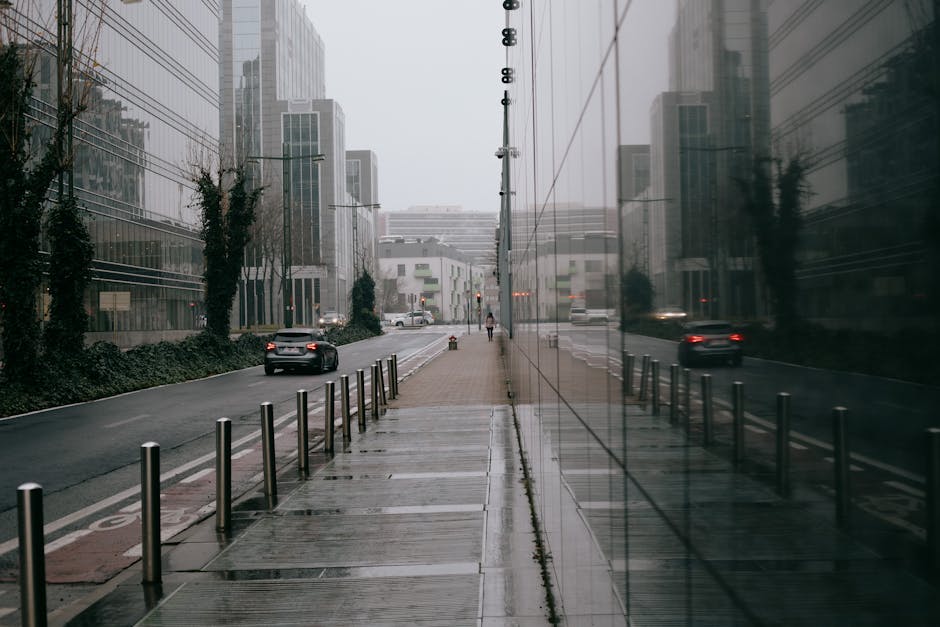 A narrow street lined with multi-storey residential buildings featuring large windows and brick facades in various colours, including grey, red, and white. The street is paved with cobblestones, and there are several parked cars, including a grey van positioned near a building's entrance, with others further down the street. A large tree with dense green foliage extends over the sidewalk, providing shade, and the overcast sky contributes to diffuse lighting. The scene captures a typical urban residential area suitable for house removals and furniture transport, illustrating the constraints of narrow streets and parking in Pentonville, which [COMPANY_NAME] navigates during home relocation and moving services, often involving loading and unloading furniture and packed boxes near the doorways and on the pavement.