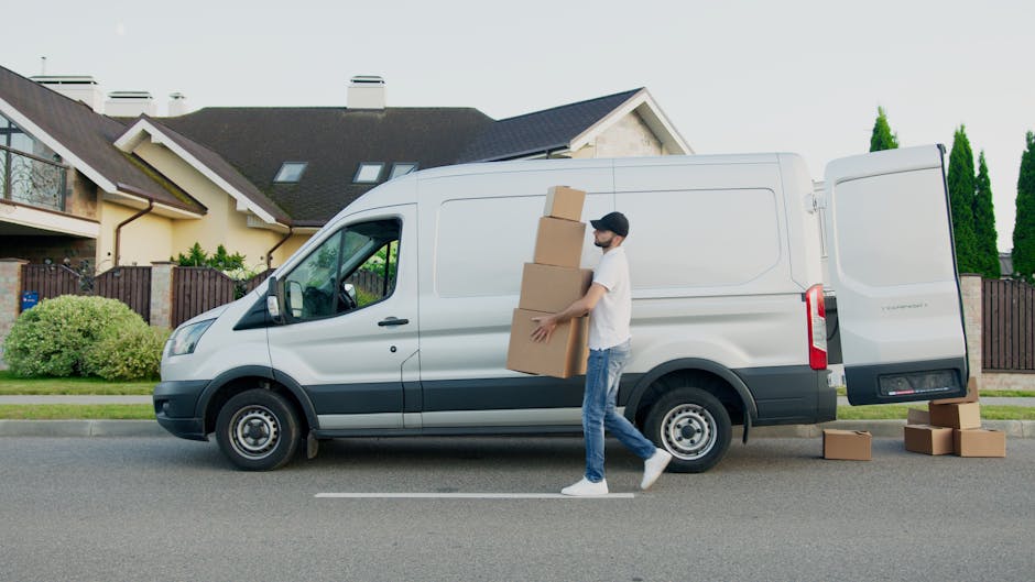 Two men are engaged in a home relocation task outside a residential property on a paved driveway. They are using a hand truck to move a large cardboard box from a white van with its rear doors open, w